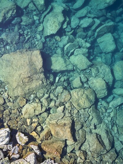 Ein klarer Bergsee in Kappl aus der Vogelperspektive. Am Grund des Sees sind große Steine und Felsen zu erkennen.