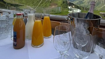 Ein Tisch mit verschiedenen Getränken, darunter Säfte und Sekt, steht vor einer beeindruckenden Berglandschaft. Die Wolken ziehen sanft über die grünen Hügel.