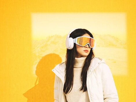 Eine Frau steht vor einer gelben Wand und trägt eine weiße Jacke sowie farbige Skibrille. Im Hintergrund ist eine verschwommene Landschaft zu sehen.