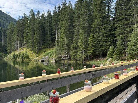 A picturesque wooden boardwalk at the shore of a clear body of water. On the boardwalk, there are glasses with colorful fruits and desserts, surrounded by tall fir trees.