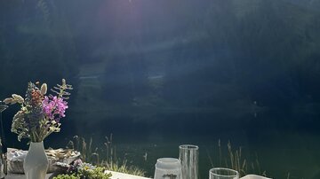 A beautifully set table with a view of a lake and surrounding forests. The sun shines brightly over the landscape.