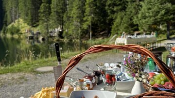 A beautifully set table in nature with fresh croissants, cheese, and fruit. In the background, tall trees and a clear blue sky can be seen.