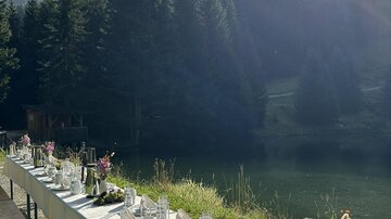 A festively set table by the edge of a pond, surrounded by tall fir trees. The sun shines over the landscape, creating a pleasant atmosphere.