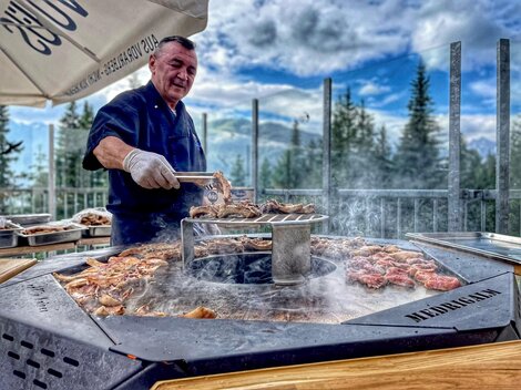 A chef is grilling meat on a round grill outdoors. In the background, trees and a cloudy sky can be seen.