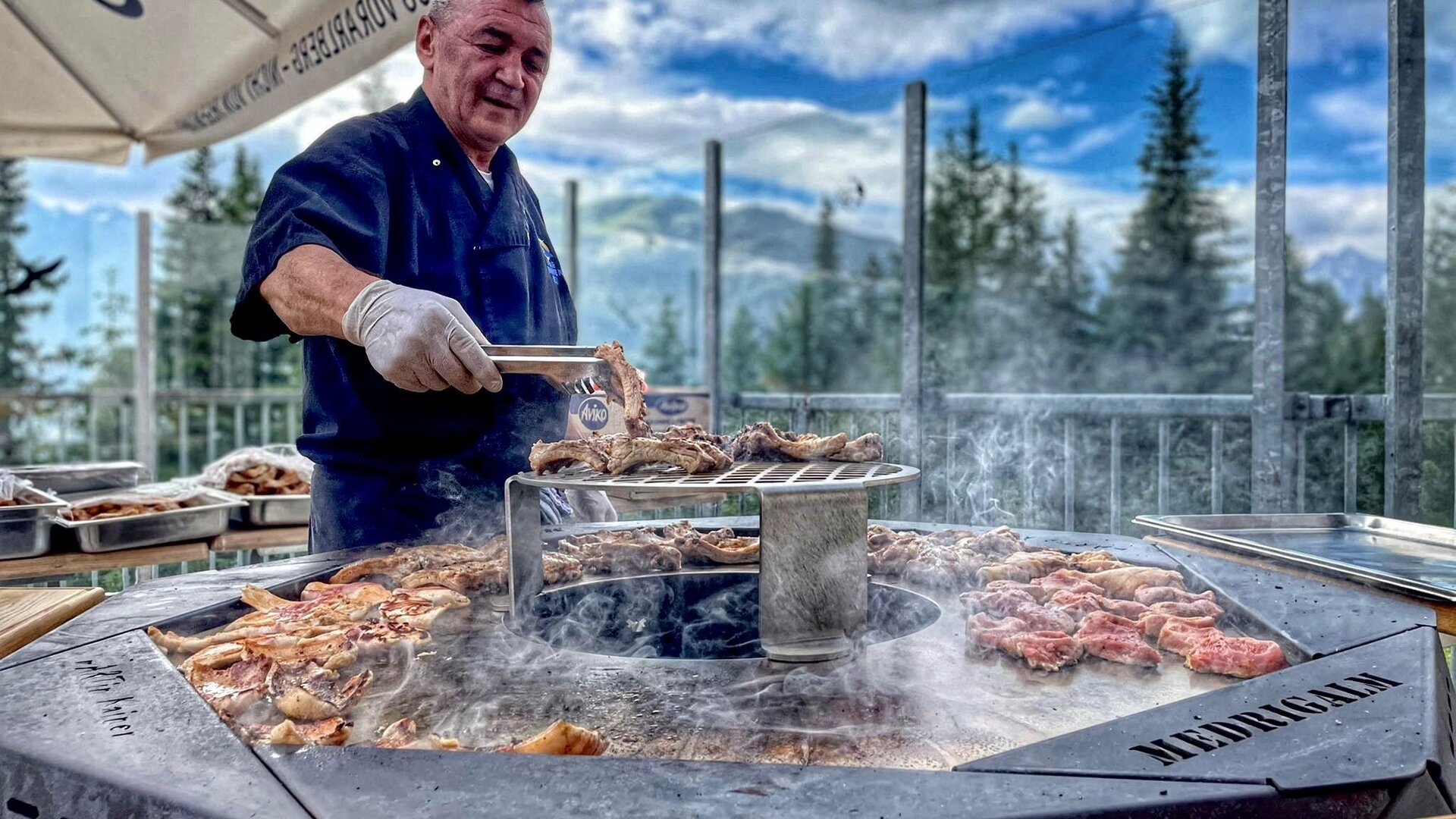 A chef is grilling meat on a round grill outdoors. In the background, trees and a cloudy sky can be seen.