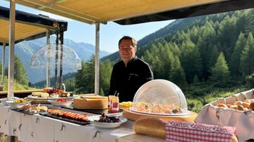 A man stands at an outdoor buffet, surrounded by a picturesque mountain landscape. The buffet is laid out with various foods and drinks.