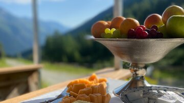 A beautifully arranged table with various types of cheese and a bowl of fresh fruit. In the background, there are mountains and a blue sky.