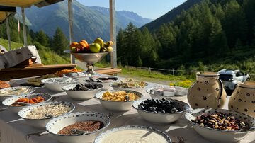 A table with various bowls full of ingredients for a buffet is set in a mountain landscape. In the background, green mountains and a clear sky can be seen.