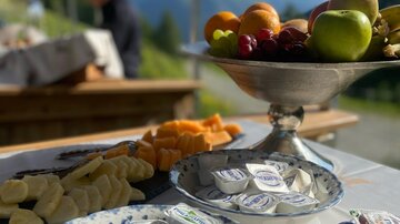 A serving table with various snacks, including fruits, cheese, and chocolates. In the background, there are mountains and a blue sky.