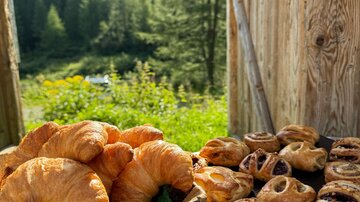 A basket with fresh croissants and a selection of pastries is on a table. In the background, green forests and mountains are visible.