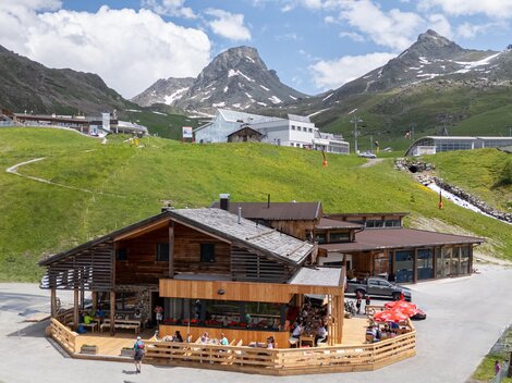 A rustic cabin in the mountains with a terrace area. In the background, green meadows and snow-capped peaks can be seen.