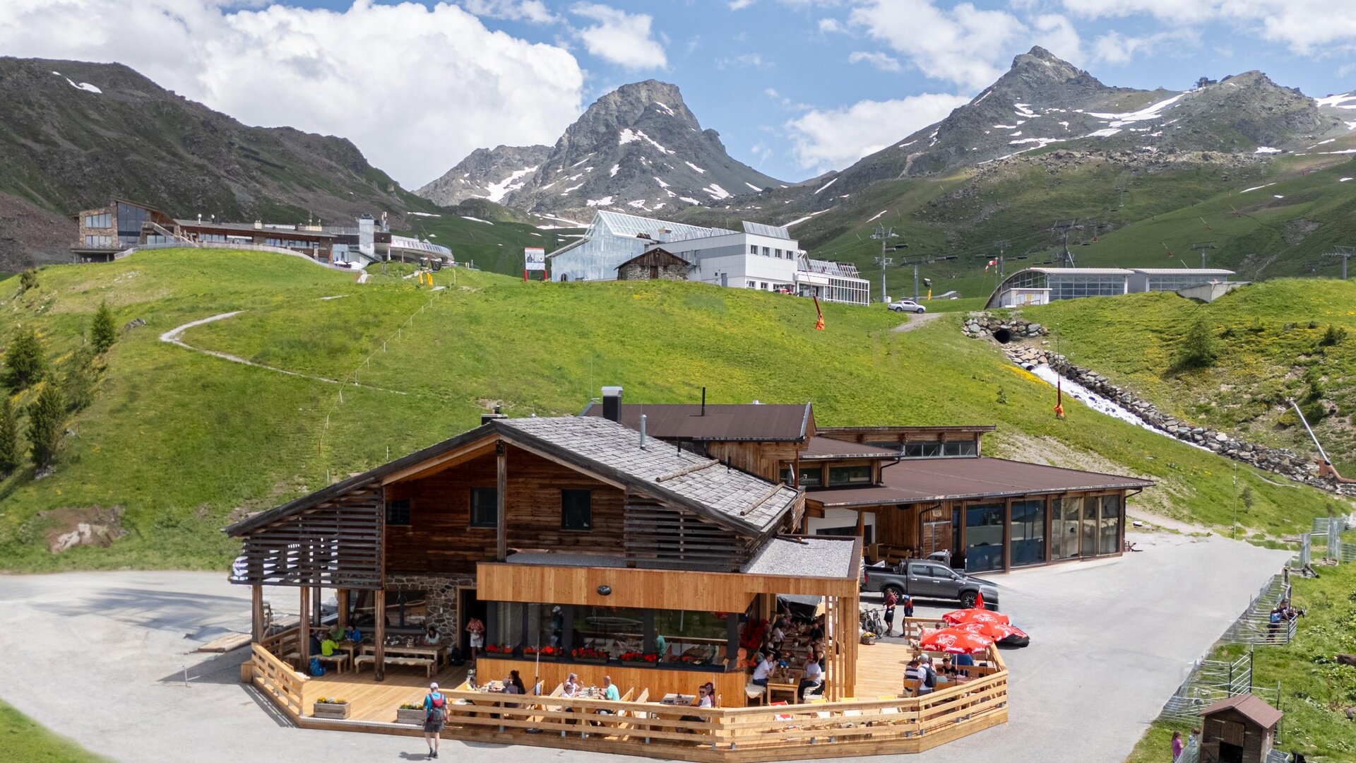 A rustic cabin in the mountains with a terrace area. In the background, green meadows and snow-capped peaks can be seen.