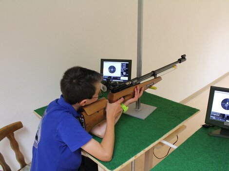 A boy is shooting at a target with an air rifle. In front of him are two monitors displaying his hits.