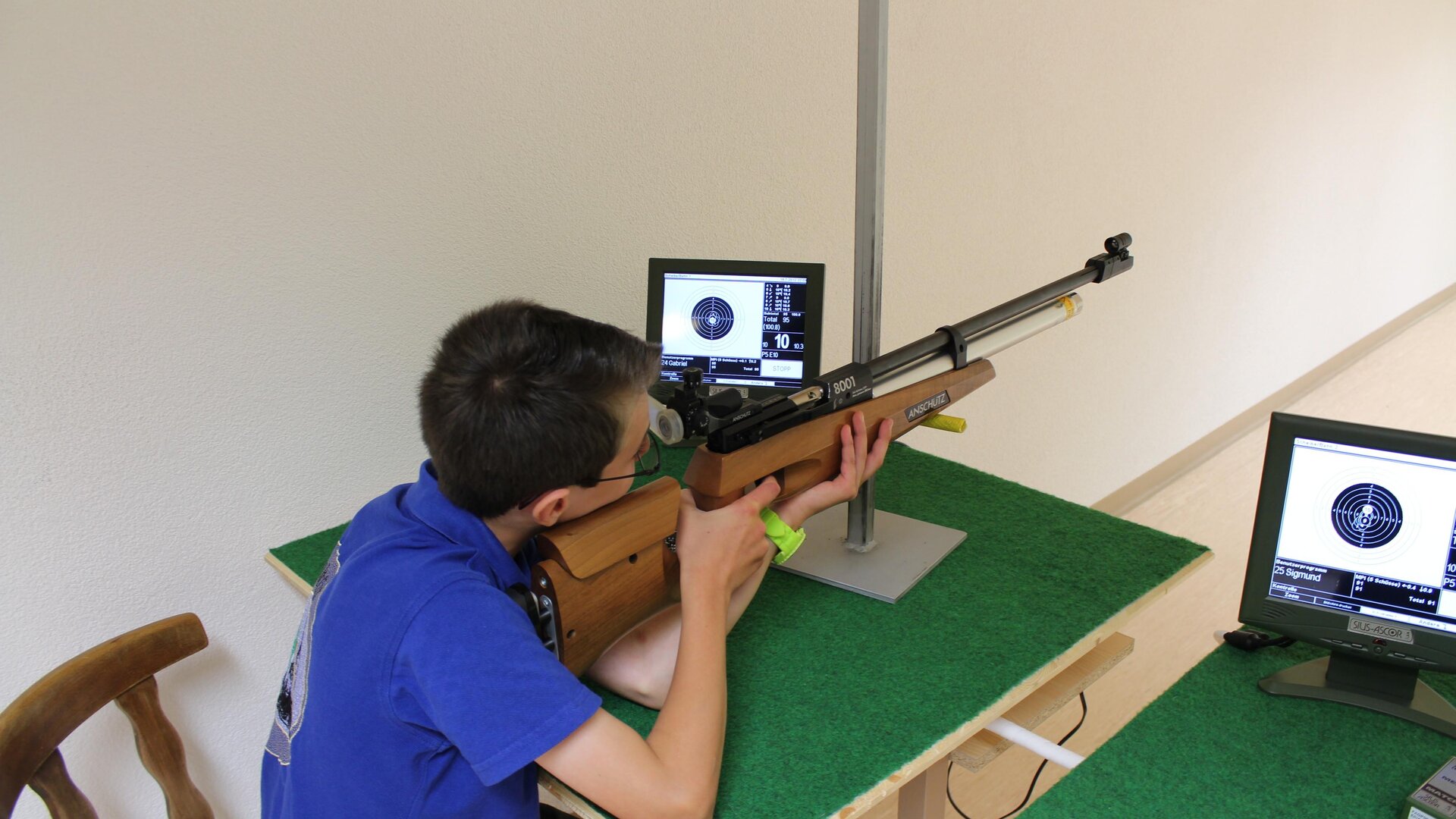 A boy is shooting at a target with an air rifle. In front of him are two monitors displaying his hits.