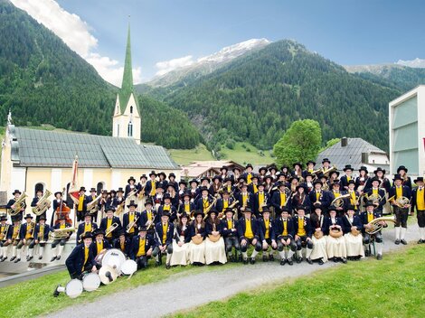 A large group of people in traditional costumes stands in a picturesque landscape. In the background, there is a church and the mountains of the Alps.