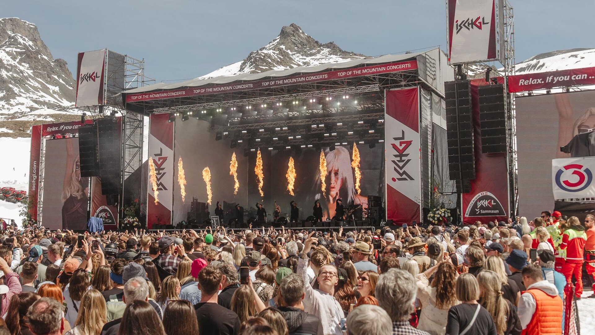 Ein großes Musikfestival in den Bergen mit einer beeindruckenden Bühne. Viele Menschen genießen die Aufführung unter klarem Himmel.