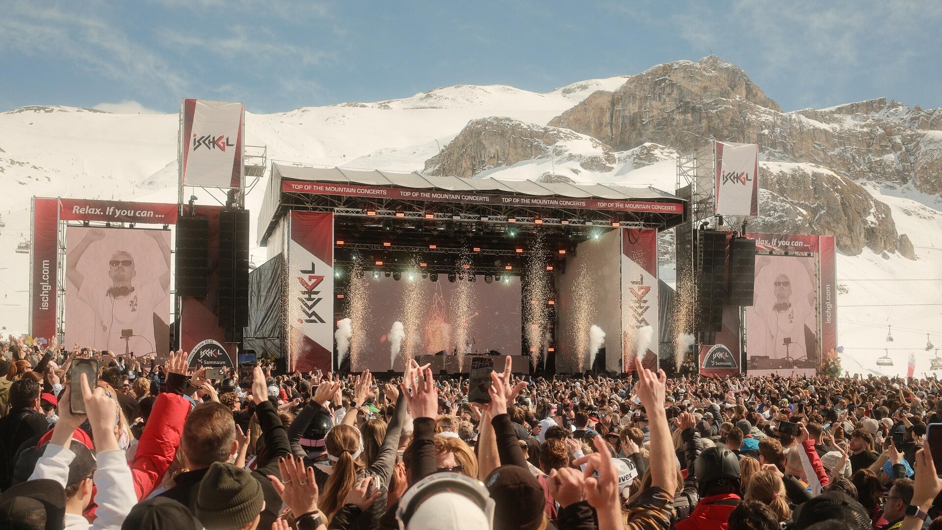 Ein großes Musikfestival in den Bergen mit vielen Menschen, die zur Show tanzen. Im Hintergrund sind schneebedeckte Berge und eine Bühne mit Lichtern zu sehen.
