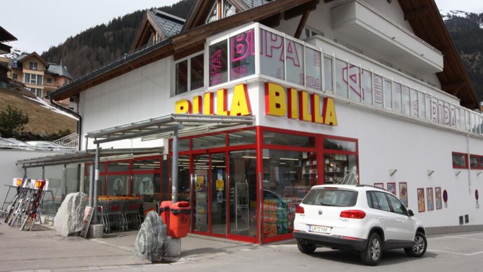 A Billa supermarket with a modern facade and large windows. In front of the store, there is a white car in a parking lot.