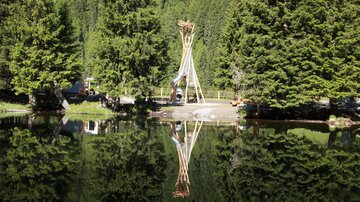 Ein schöner Teich umgeben von hohen, grünen Bäumen. Im Vordergrund ist eine Holzskulptur zu sehen, die sich im Wasser spiegelt.