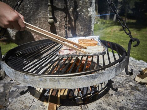 Ein Grill mit rauchenden Würstchen wird über einer offenen Flamme zubereitet. Im Hintergrund ist das grüne Gras und die Natur zu sehen.