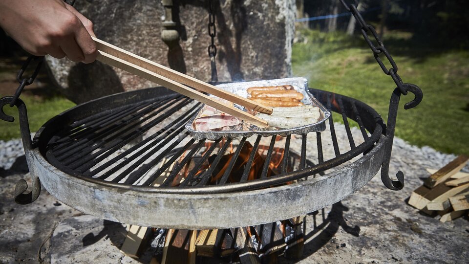 Ein Grill mit rauchenden Würstchen wird über einer offenen Flamme zubereitet. Im Hintergrund ist das grüne Gras und die Natur zu sehen.