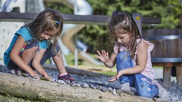 Zwei Kinder spielen im Freien und sammeln Steine auf einem Baumstamm. Im Hintergrund sind ein Spielplatz und grüne Wiesen zu sehen.