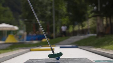 A mini golf course with a putter and a target area is in the foreground. In the background, there are playground equipment and green meadows.