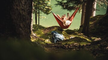 Eine Person entspannt in einer Hängematte zwischen Bäumen am Ufer eines Sees. Neben der Hängematte liegt ein Rucksack auf dem moosbedeckten Boden.