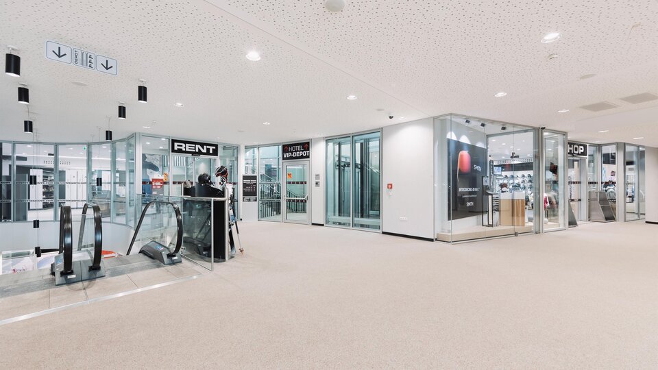 A modern interior with windows and shops. On the left side, there is an escalator and a reception desk.