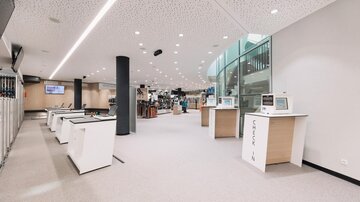A modern, bright interior with multiple reception desks. In the background, fitness equipment and a spacious area are visible.