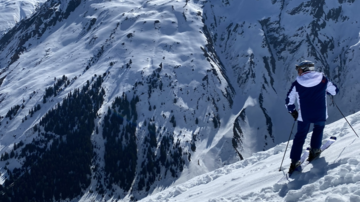 Ein Skifahrer steht auf einer verschneiten Piste mit hohen Bergen im Hintergrund. Der Himmel ist klar und blau.