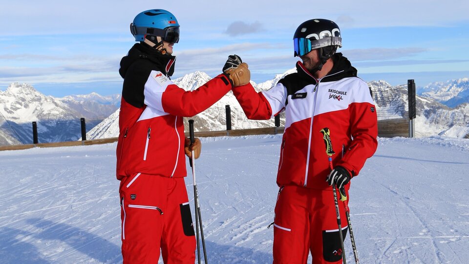 Two skiers in red ski suits stand on a snow-covered peak. Majestic mountains can be seen in the background.