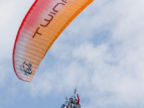Ein Gleitschirmflieger schwebt hoch am Himmel. Der Schirm ist orange und rot, und die Wolken sind leicht sichtbar.