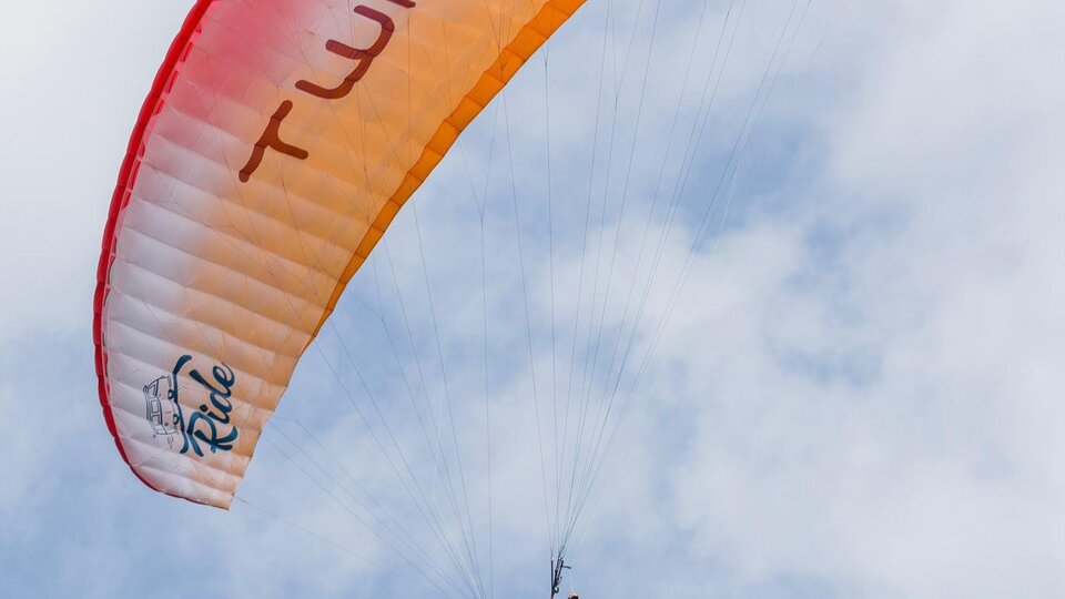 Ein Gleitschirmflieger schwebt hoch am Himmel. Der Schirm ist orange und rot, und die Wolken sind leicht sichtbar.