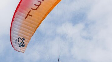 Ein Gleitschirmflieger schwebt hoch am Himmel. Der Schirm ist orange und rot, und die Wolken sind leicht sichtbar.
