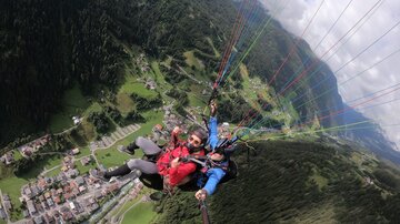 Drei Personen, die beim Paragliding über eine grüne Landschaft fliegen. Im Hintergrund sind Berge und ein Dorf zu sehen.
