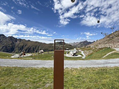 Eine Berglandschaft mit Seilbahnen und einem klaren blauen Himmel. Im Vordergrund steht ein rechteckiger Pfosten vor einer grünen Wiese.