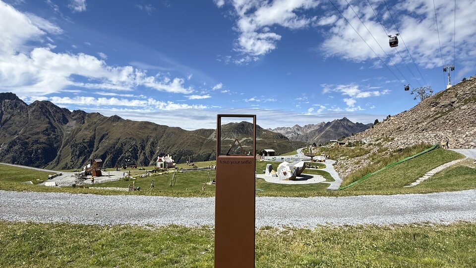 Eine Berglandschaft mit Seilbahnen und einem klaren blauen Himmel. Im Vordergrund steht ein rechteckiger Pfosten vor einer grünen Wiese.